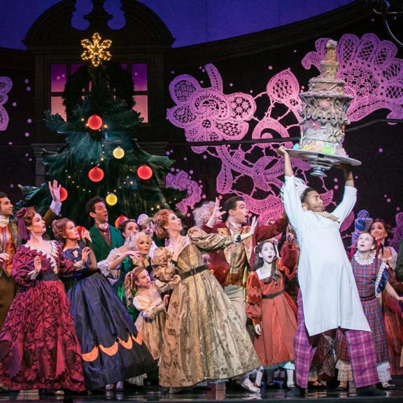 Various Dancers in costumes staring at a baker holding a cake high above his head