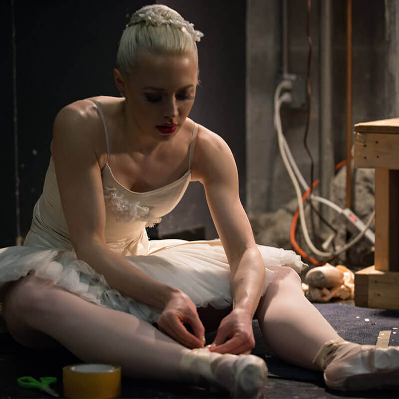 Dancer typing pointe shoes while seated on stage 