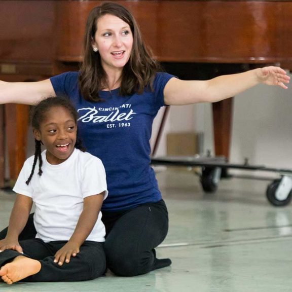 Photo of a female ballet teacher wearing a Cincinnati Ballet showing a young child how to pose with their arms