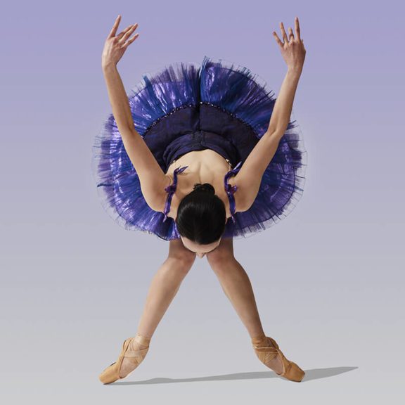 Cincinnati Ballet Ballerina In A Purple Tutu, Bent Over With Her Arms Straight In The Air Behind Her