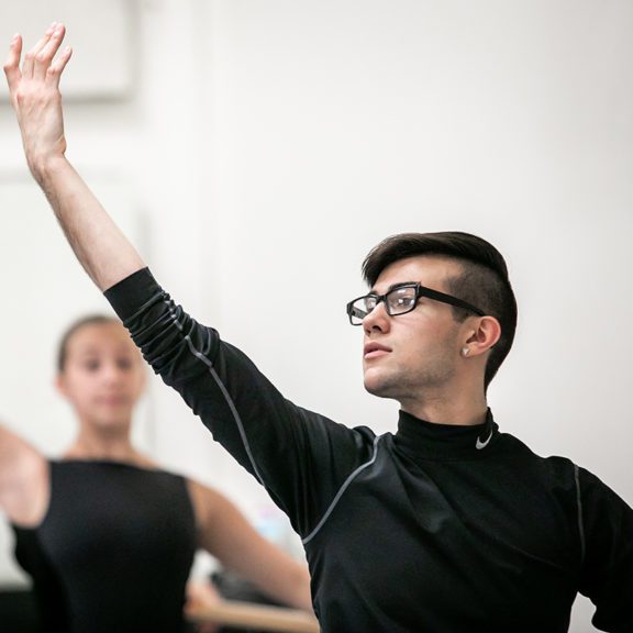 Photo of a male ballet dancer wearing a black Nike shirt and black glasses as he poses with his arm in the air