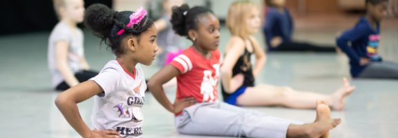 Photo of young girls sitting on the ground with their hands on their hips during a Cincy Dance class
