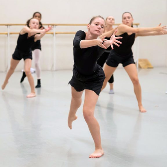 Photo of five female ballet dancers in black outfits reaching out to their left in a pose