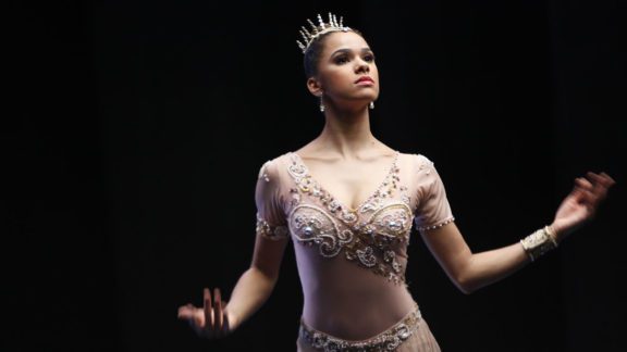 Photo of a ballerina posing and wearing a dance costume with a crown on her head with a black background behind her
