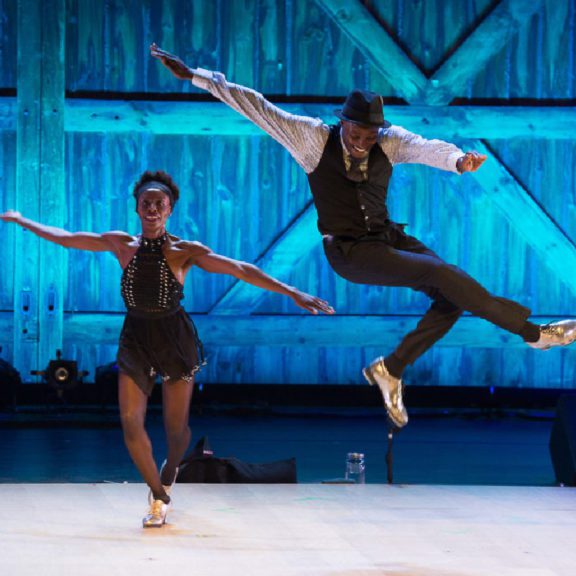 Photo of a male ballet dancer jumping and posing in the air with a female ballerina dancing on the floor beside him