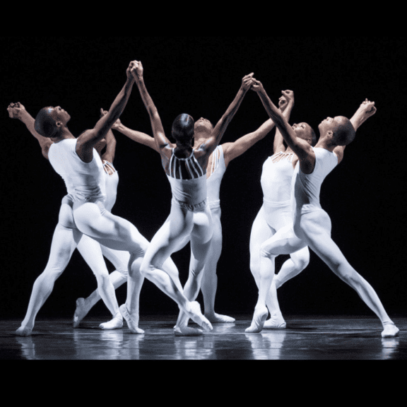 Photo of ballet dancers in white leotards posing with their arms in the air together during a performance
