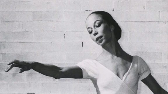 Black and white photo of a ballerina standing in front of a white brick wall