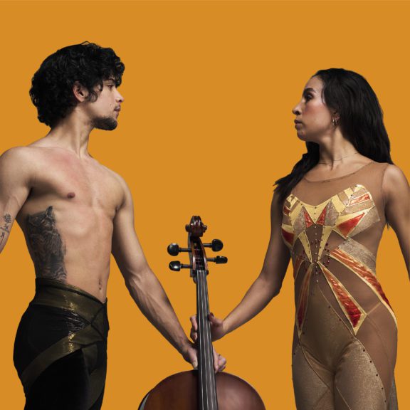 Photo of a male ballet dancer and female ballet dancer both holding onto a musical instrument as they stare at each other with a gold background behind them