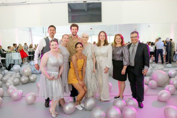 Photo of men and women standing together for a picture in a room full of balloons on the floor and a mirror behind them