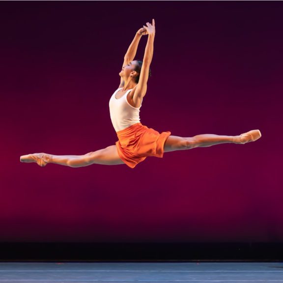 Photo of a ballet dance leaping through the air with their legs stretched into a split and their arms straight above their head