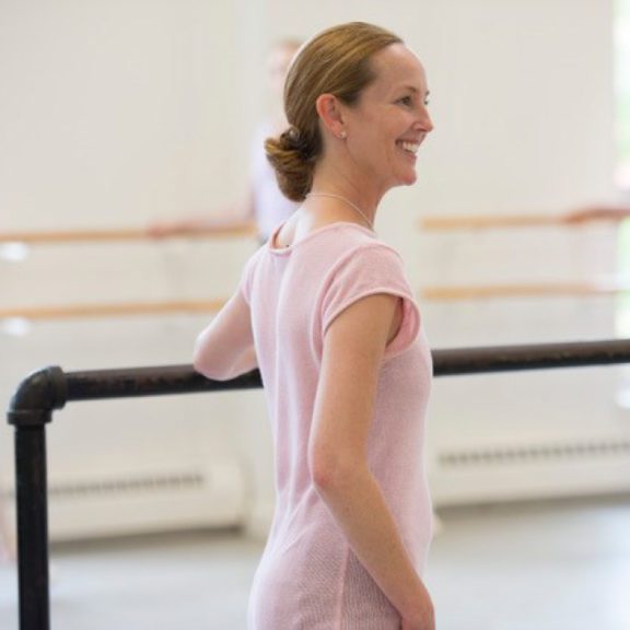 Photo of a woman in a pink dress smiling as she holds onto a balance bar in a ballet dance studio while she teaches