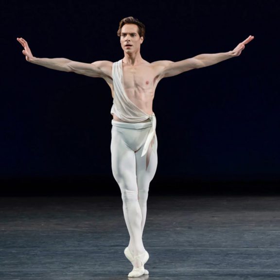 Brunette male ballet dancer wearing white tights and a white tied shirt around him as he poses with his arms out