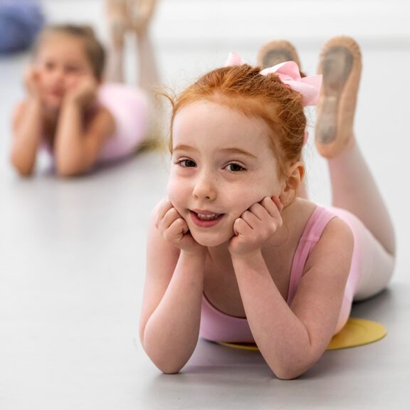 Photo of two small female ballerinas laying on the ground with their hands on their cheeks and their feet up toward their heads