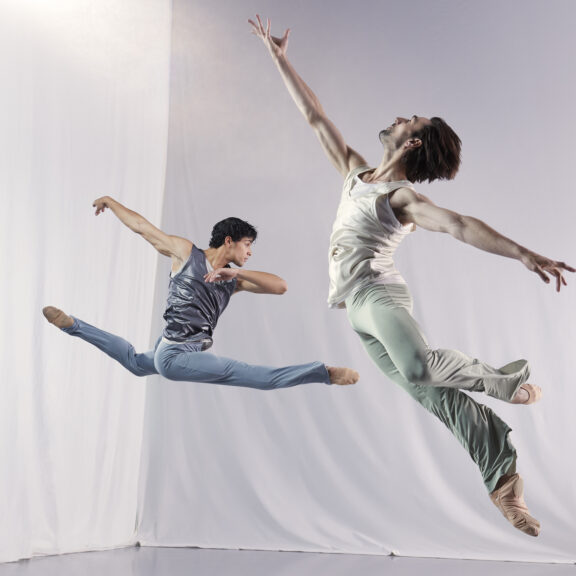 2023-2024 season | Photo of two male ballet dancers leaping through the air with a white curtain behind them