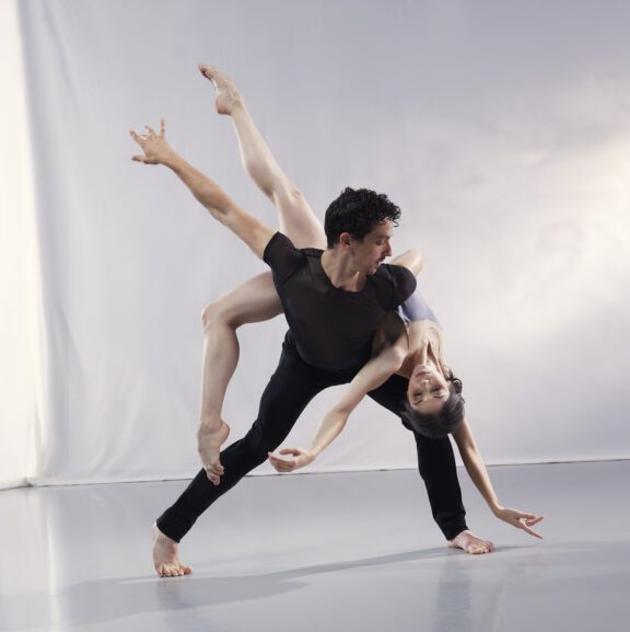2023-2024 season | Photo of a male ballet dancer wearing a black shirt and black pants holding a female ballerina in a purple leotard on his hip as she poses upside down