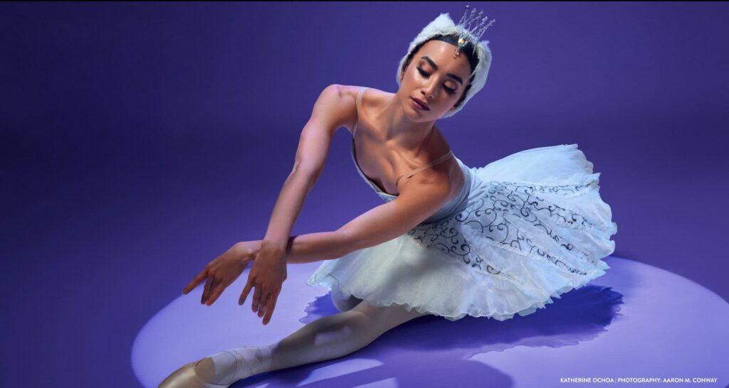 Ballet dancer in a white Swan Lake costume kneels gracefully on a purple studio backdrop, arms extended forward and head bowed in a poised, elegant pose.
